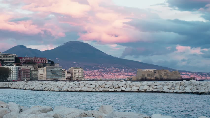 Mount Vesuvius towers over Naples Bay at dusk, with Castel dell’Ovo, white breakwater rocks, calm water, and pink storm clouds above the waterfront.