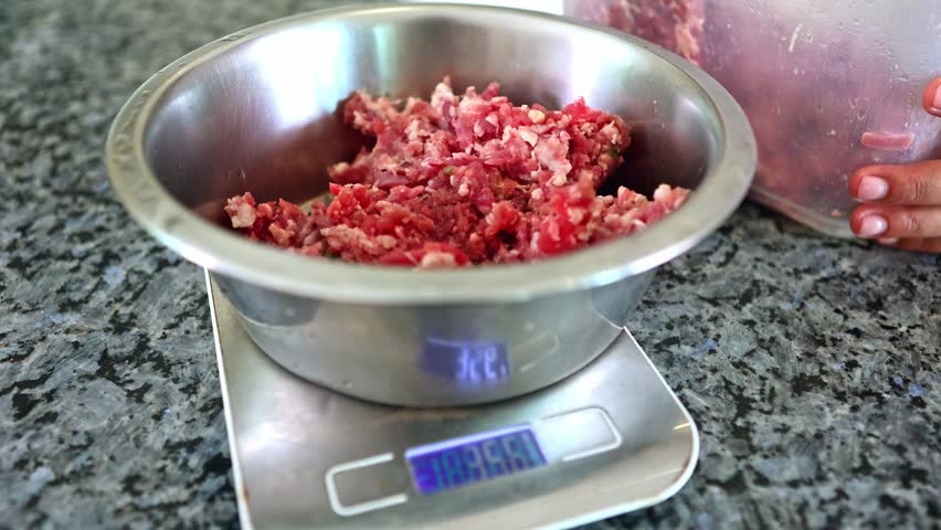 Close up of a person using a digital kitchen scale to weigh the correct portion of minced raw meat into a stainless steel bowl for a biologically appropriate raw food diet for a pet