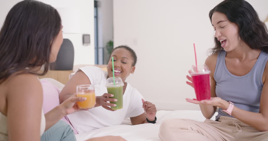 Diverse women friends on bed in bright room, center offering green cup, toasting red-orange drinks. Trio, colorful, gathering, conversation, loungewear, cozy, casual