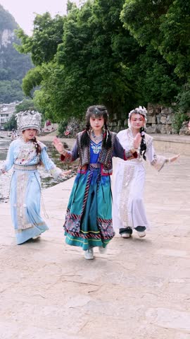 Three beautiful young Chinese women performing a traditional folk dance by a river in the scenic landscape of Yangshuo, dressed in colorful ethnic costumes with ornate headdresses