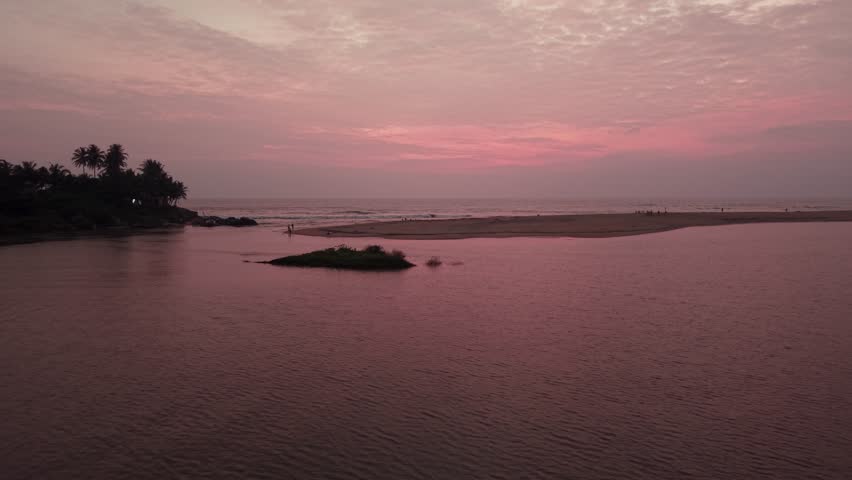Aerial view of a calm river mouth meeting the ocean under a vibrant pink sunset sky with palm trees and a small sandbar in Galle, Southern Province, Sri Lanka.