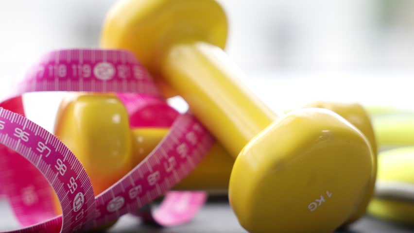 Measuring tape and dumbbells on blurred background, closeup. Camera moving left