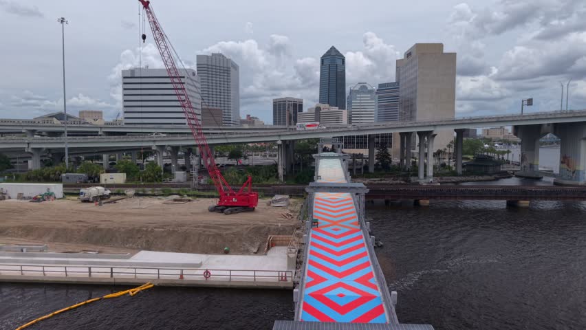 Aerial view of a construction site with a red crane and a colorful chevron-patterned bridge near the river and city skyline in Jacksonville, Florida, United States.