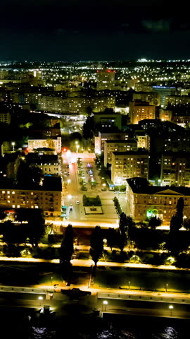 Vertical video. Saratov, Russia. Oktyabrskaya Street runs through the heart of the city as a broad, tree-lined avenue flanked by mid-rise residential blocks and administrative buildings, all glowing with warm window light against the dark sky. The footage captures a smooth aerial pullback that gradually reveals the full scale of the urban grid, with the street acting as a central axis guiding the eye toward the distant horizon. The architecture is predominantly Soviet-era panel construction, interspersed with green courtyards and pedestrian walkways. As the camera ascends, the Volga River embankment comes into view at the lower edge of the frame, its promenade lights tracing the waterfront. The scene is set deep in the night, under a partly cloudy sky, with the amber and white illumination of streetlamps creating strong contrast against the dark foliage and building silhouettes. The overall atmosphere is calm and expansive, offering a sweeping view of Saratov