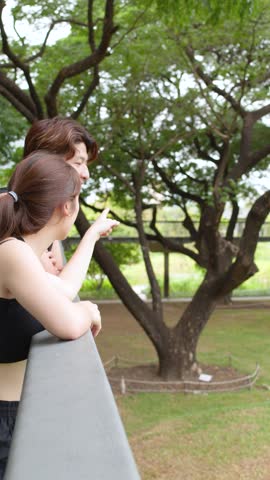 A young couple in black athletic wear stands on a bridge, pointing at park scenery
