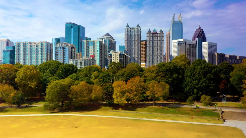 Dynamic drone footage soaring above Piedmont Park showing lush green trees transitioning to autumn colors with majestic skyscrapers of Midtown Atlanta under blue sky and soft white clouds.