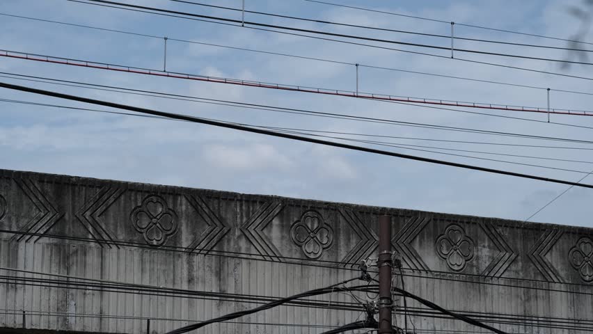 Modern commuter train traveling on elevated railway bridge with overhead wires, concrete barrier, and urban transportation infrastructure.