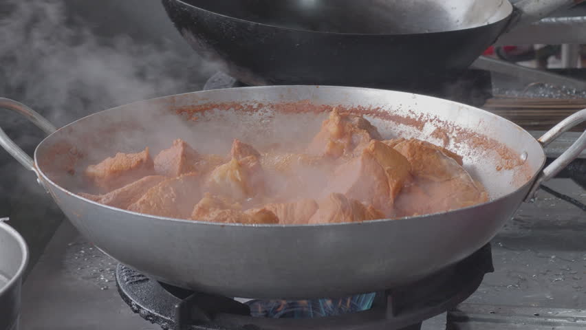 Large chunks of seasoned pork meat simmering and boiling in a large wok on a gas stove with steam rising, close up of traditional street food cooking