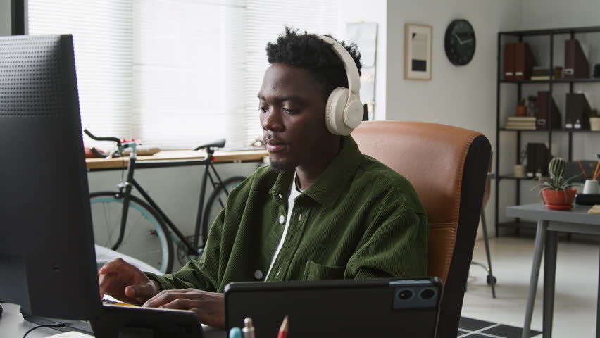 Medium side shot of focused young African American male programmer writing code for mobile app on desktop computer in startup office, listening to music, smartphone, tablet and laptop on desk
