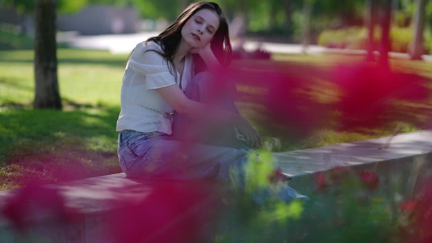 Pensive young woman sitting alone on a park bench, looking away with a thoughtful expression. Beautiful sunlit scene with blurry red flowers