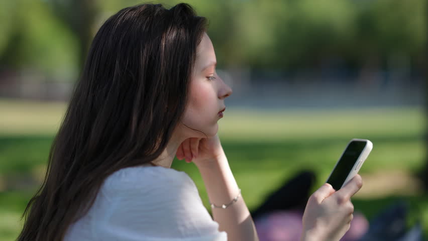 Profile view of a thoughtful young woman with long hair sitting outdoors on a sunny day. She is holding and looking at her cellphone