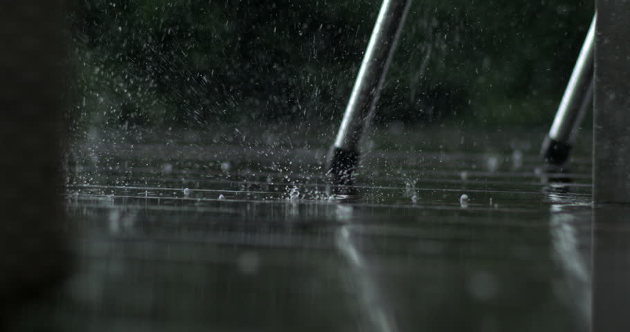 Close up of rain drops bouncing off wet wood near metal chair leg, recorded in super slow motion with high speed camera, garden scene in background