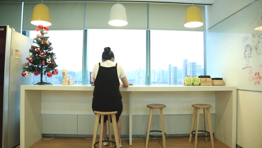 A rear-view shot of an individual sitting at a bright, window-side counter in a modern, clean space, likely an office or study area. A decorated Christmas tree stands to the left, and small plush toys are placed on the counter to the right. The peaceful, focused atmosphere highlights a productive and warm work or study environment.