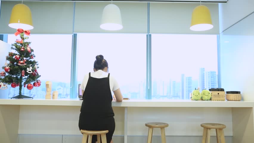 A rear-view shot of an individual sitting at a bright, window-side counter in a modern, clean space, likely an office or study area. A decorated Christmas tree stands to the left, and small plush toys are placed on the counter to the right. The peaceful, focused atmosphere highlights a productive and warm work or study environment.