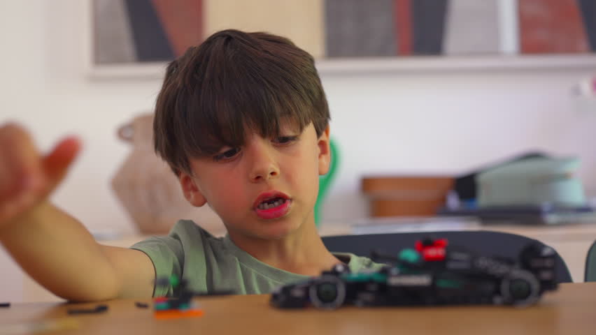 Boy examines underside of toy race car while adjusting block pieces, focused on structure and small mechanical details at table indoors