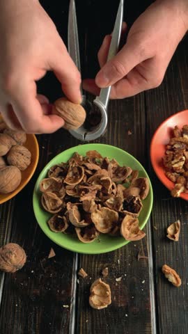 Hands using a nutcracker to break walnut shells, revealing the nut within, with empty shells and whole walnuts arranged on a wooden table, creating a rich visual of the process