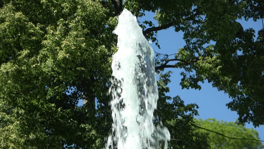 Powerful stream of water from a fountain gushing upwards against trees and a clear blue sky