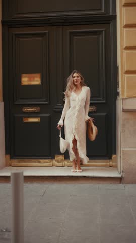 Beautiful young blonde woman in a white dress and straw hat dancing joyfully on the steps of a classic parisian building, smiling and having fun during her romantic vacation in france