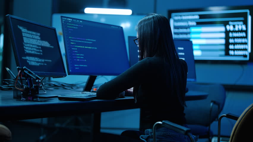 Focused female programmer coding on multiple monitors in a dark high-tech cybersecurity center. IT specialist typing on keyboard, analyzing big data on screens in a server room late at night.