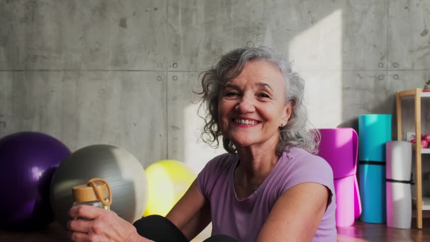Elderly woman with grey hair smiling at the camera, holding a water bottle after a home workout