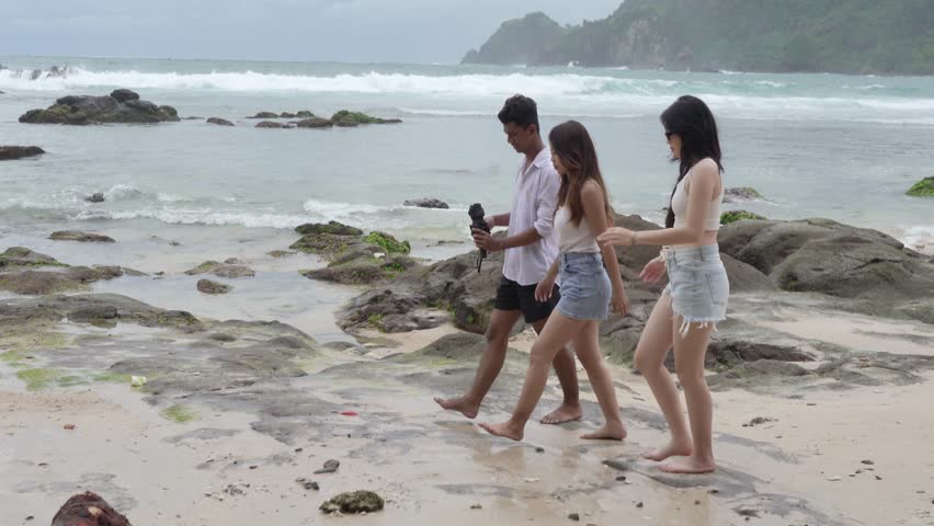 Three young friends walking barefoot along the shoreline of Wedi Ombo Beach, Yogyakarta, enjoying a tropical vacation and coastal lifestyle in Indonesia.