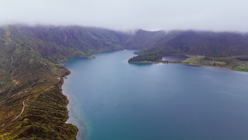 Crater lake in volcanic terrain surrounded by lush greenery and cloud cover. Natural wilderness of Azores, Portugal, drone video