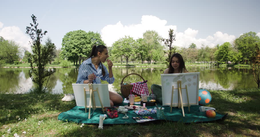 Two friends enjoy a relaxing day painting by the lake. They share snacks and drinks, surrounded by nature