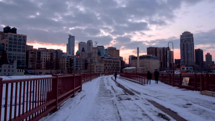 Cinematic motion shot of city skyline during winter sunset with snow on bridge. Pedestrians walk along historic walkway while urban lights begin to glow under colorful evening clouds in city.