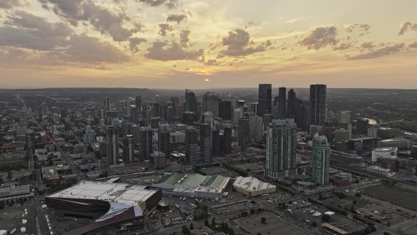 Cinematic drone flight over Calgary Alberta at sunset showing high rise buildings and skyscraper architecture under orange sky with clouds. Dynamic city landscape at dusk with urban horizon.