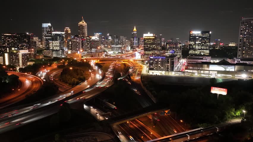 Aerial view of Atlanta Georgia city skyline at night showing heavy vehicle traffic on freeway interchange. Cinematic drone shot captures downtown buildings and glowing street lights in motion.