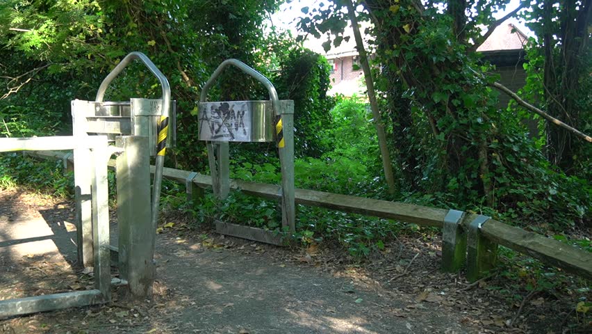 Entrance of a public park footpath featuring a pair of heavy-duty galvanized metal A-frame kissing gates or motorcycle inhibitors, surrounded by green trees and a wooden fence.
