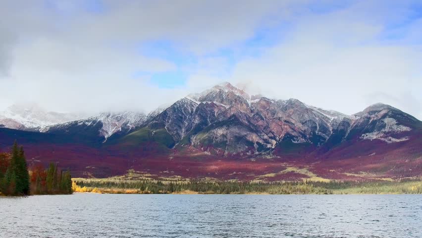 Banff. A serene landscape photograph featuring a mountain range with snowcapped peaks, a body of water, and a forested area. The sky is partly cloudy, with a mix of blue and white hues.