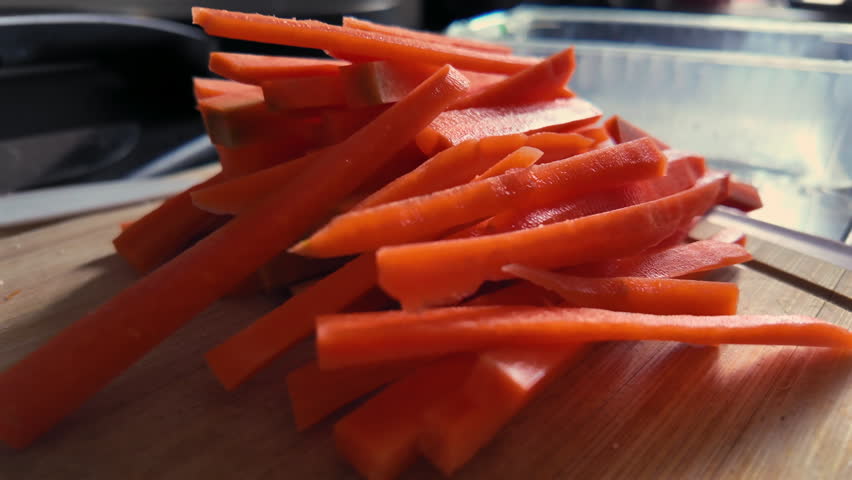 Fresh carrot sticks chopped on wooden cutting board beside raw chicken, ready for meal preparation in home kitchen with morning light and soft shadows