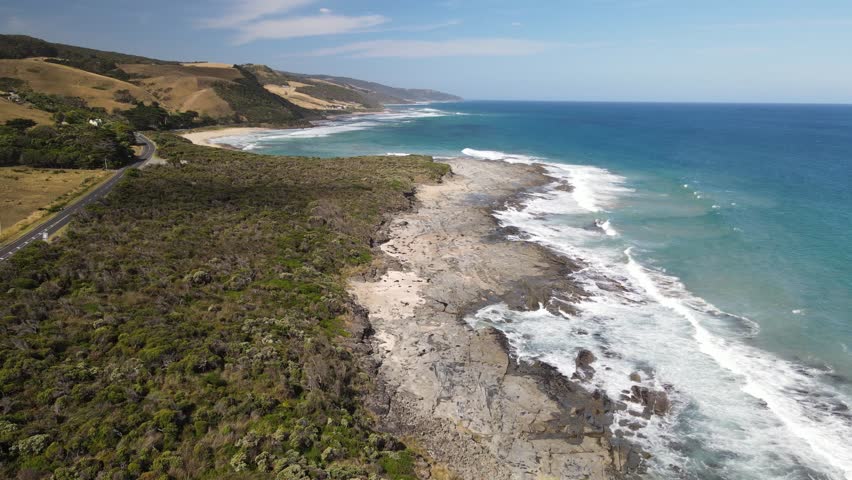 Beautiful panoramic aerial view of a scenic coastal road winding near a rugged rocky ocean coastline with vibrant turquoise waves, Great Ocean Road, Victoria, Australia