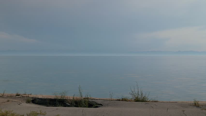 A handheld static shot capturing the quiet, smooth surface of Lake Baikal in Russia. The hazy horizon features distant mountains under an overcast sky, viewed from a concrete shore with sparse weeds.