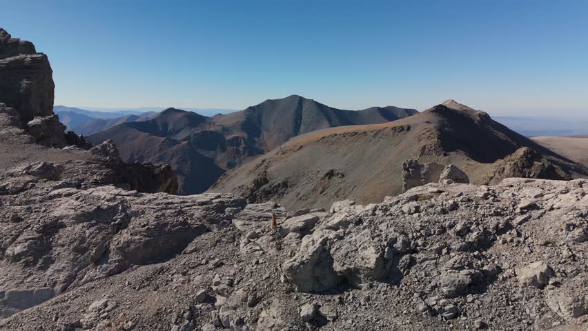 Drone footage captures mountain goats bounding along a barren ridge through harsh alpine terrain, with dramatic rocky cliffs, steep slopes and clear blue sky above distant mountain peaks