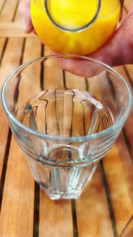 Vertical video - Closeup slow motion shot of hands pouring fresh cold orange juice from a bottle into a tall clear glass tumbler, outside on a wooden table.