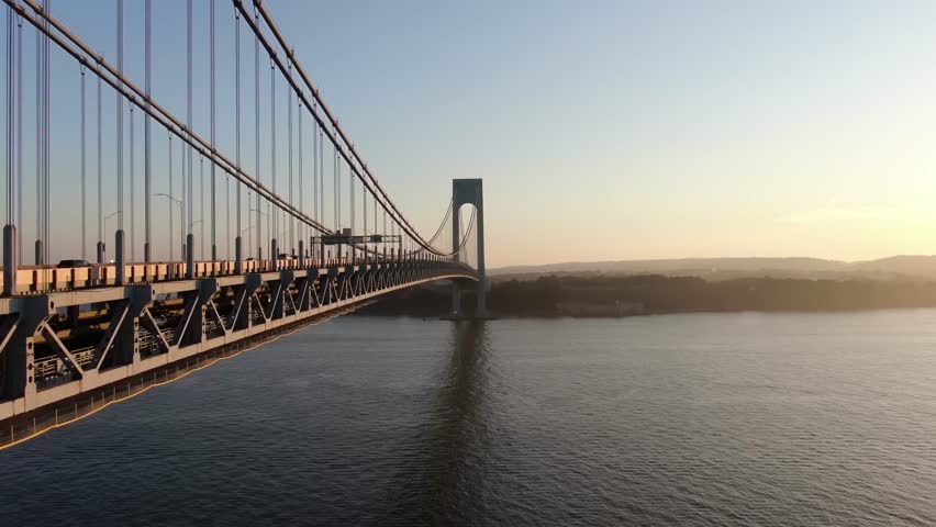 Cinematic drone footage of a suspension bridge stretching across calm water during sunset.