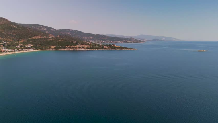 A rising aerial drone shot over the blue Aegean Sea towards the mountainous coastline and emerald hills of Kavala during a bright summer day.