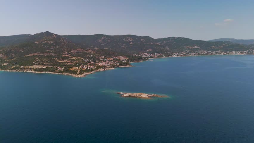 Forward-moving aerial drone shot over the calm blue Aegean Sea approaching the rugged green mountains and rocky shoreline of Kavala, Greece.