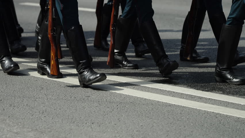 Close-up view of uniformed soldiers marching in formation along an asphalt street, black boots stepping in unison with wooden-stock rifles held at their sides during a parade.