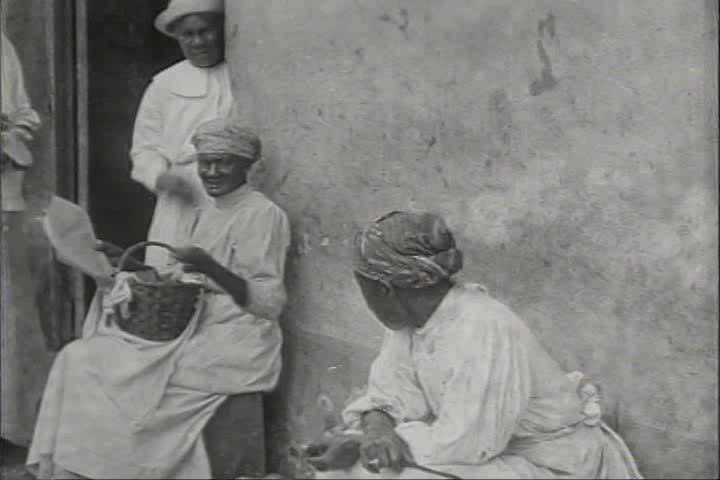 1910s, 1920s - Scenes of women of African decent conversing outside a home. A woman with a head wrap chews and looks at the camera.