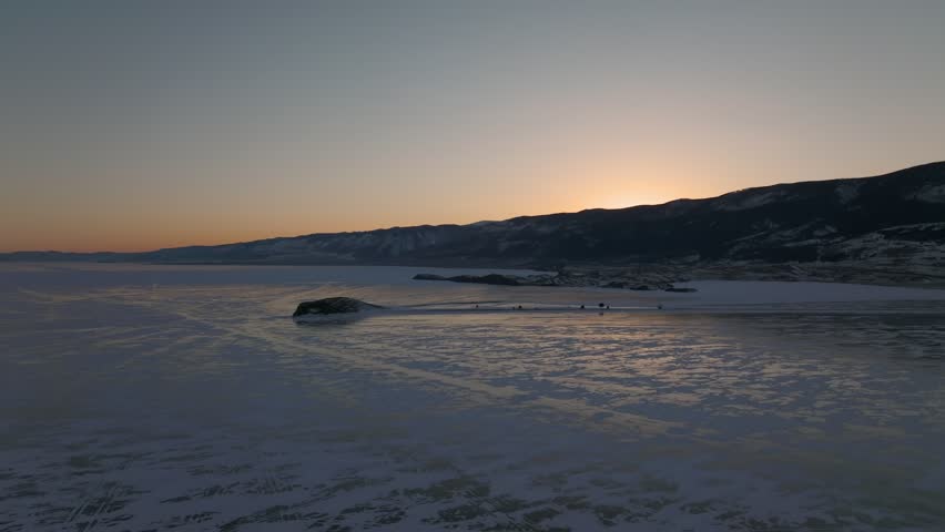 Aerial footage of frozen Lake Baikal at sunset, with cracked ice, snowy shoreline and dark mountain ridge on the horizon, calm winter landscape in Russia