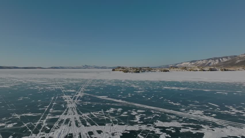 Drone footage of a vast frozen surface of Lake Baikal with rocky islets, ice patterns, and snow-covered coast under clear sky, Siberia, Russia
