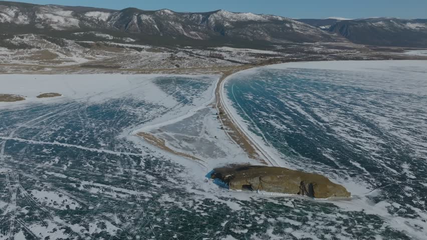 Drone footage of frozen Lake Baikal with blue cracked ice, snowy shoreline and a narrow road, with mountain range in the background, Siberia, Russia