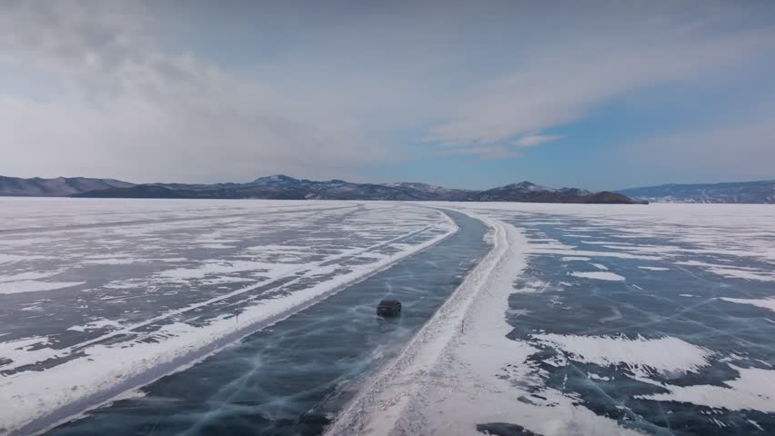 Drone footage of a vehicle driving on an ice road across frozen Lake Baikal in winter, surrounded by snow and blue ice with distant mountains, Siberia, Russia