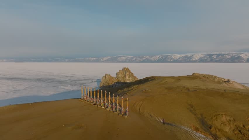 Drone footage of Shamanka Rock on Olkhon Island surrounded by frozen Lake Baikal, Irkutsk Oblast, Russia, with snowy mountains on the horizon