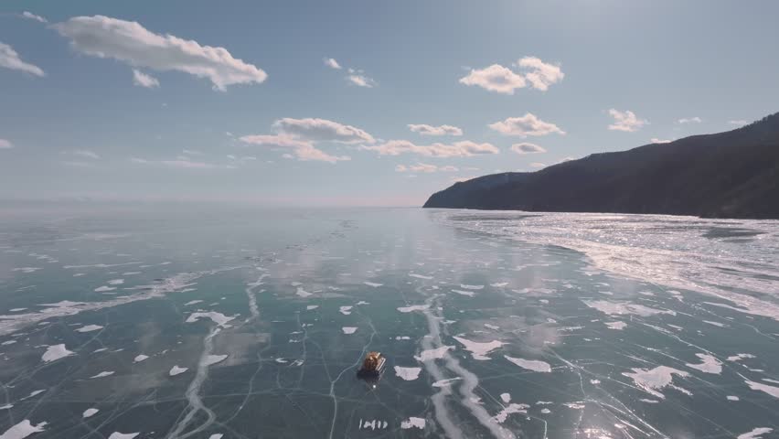 Aerial winter footage of a hovercraft traveling across frozen Lake Baikal with clear ice, cracks and snow patches, mountains on the shore, Irkutsk Oblast, Russia