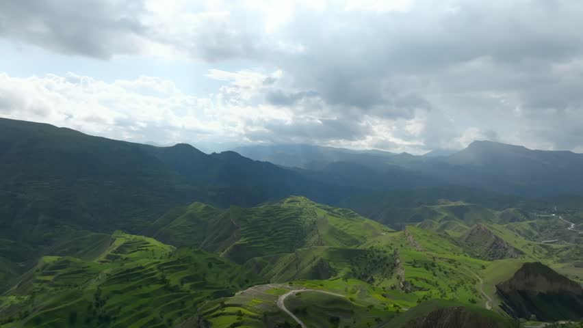 Wide aerial shot of the Chokh Terraces, terraced green hills and mountain ridges under dramatic cloudy sky in Dagestan, Russia, scenic rural nature and travel view