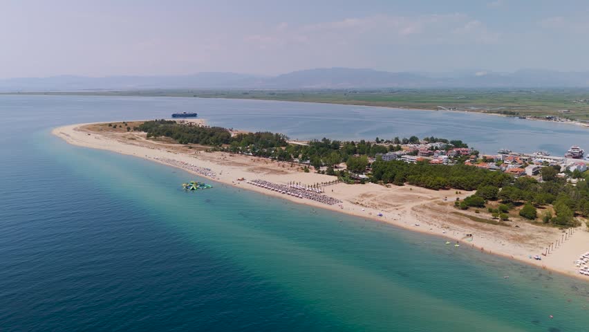High-angle drone shot descending over the turquoise Thracian Sea towards the white sand shoreline and lush green pine forest of Keramoti Beach in Northern Greece.